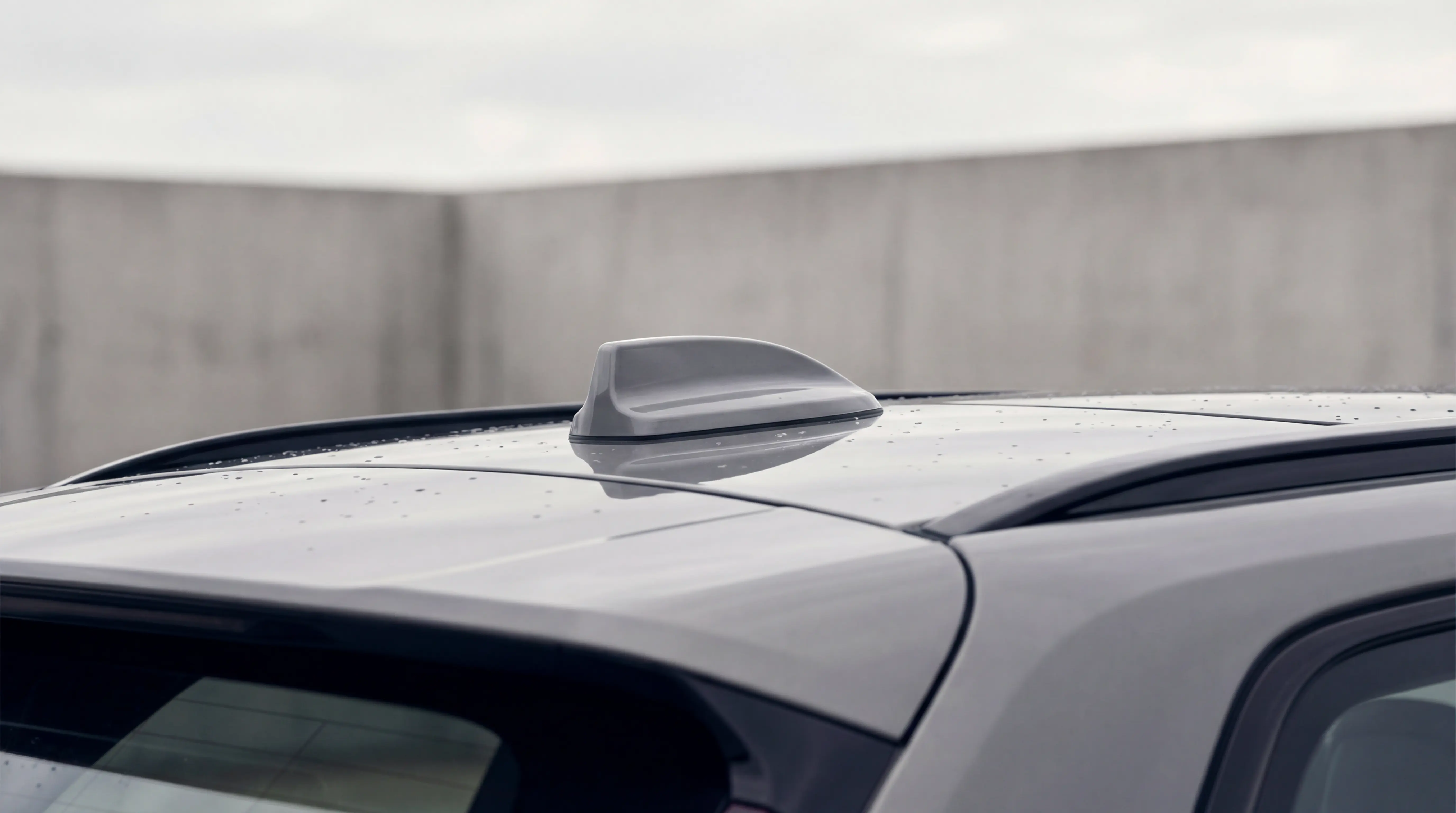 Close-up of a gray car roof with a shark fin antenna and water droplets on a cloudy day.