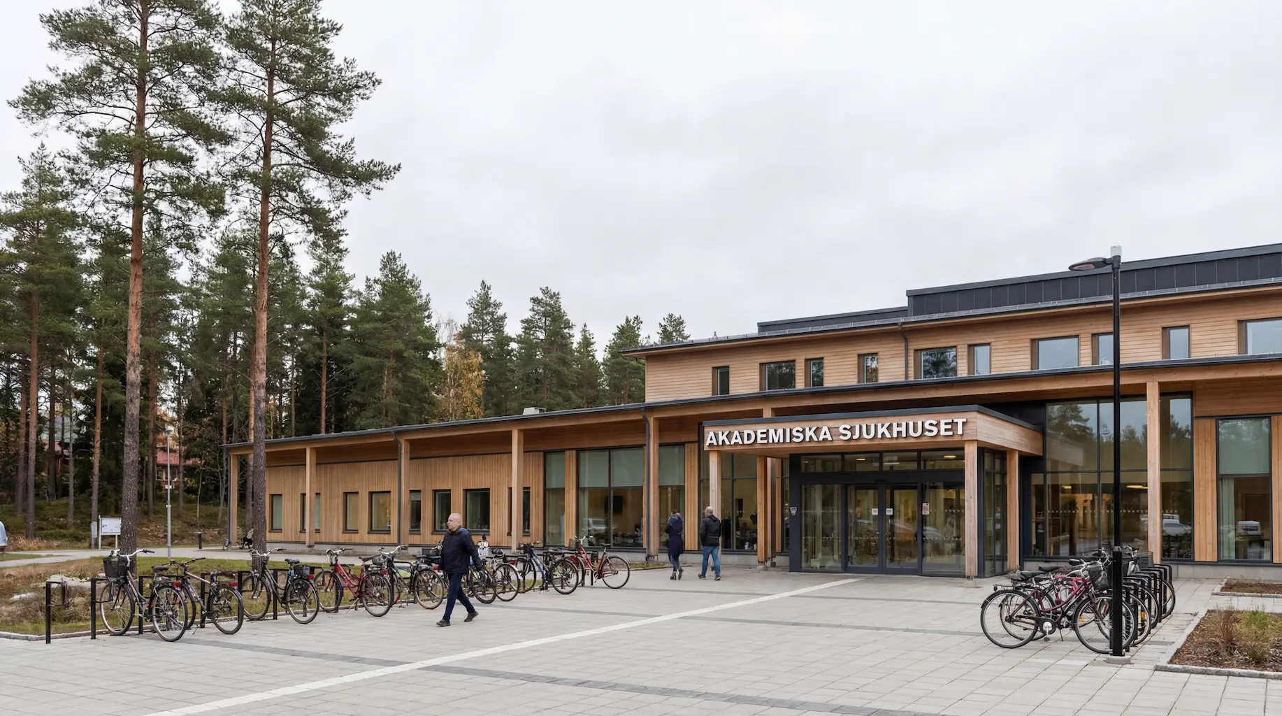 Modern wooden building of Akademiska Sjukhuset with bicycles parked outside and people walking near the entrance.